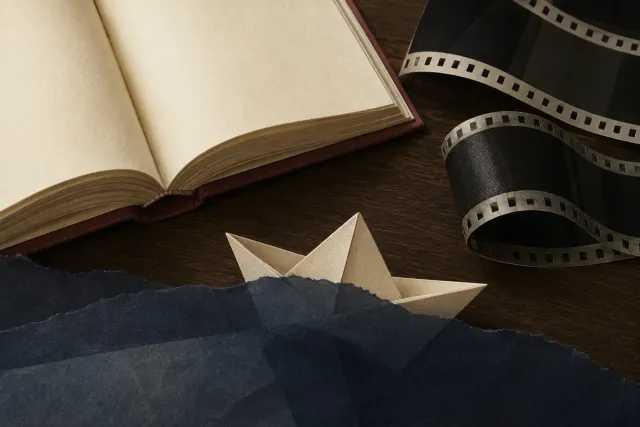 An open book and blank filmstrip lie beside a paper boat partly concealed by dark paper.